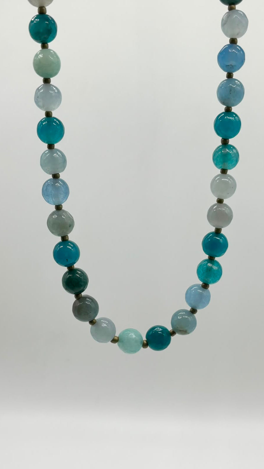 A beaded necklace made of polished round gemstone beads in shades of teal, blue, green, and pale aqua, separated by small dark spacer beads, shown against a plain light gray background.