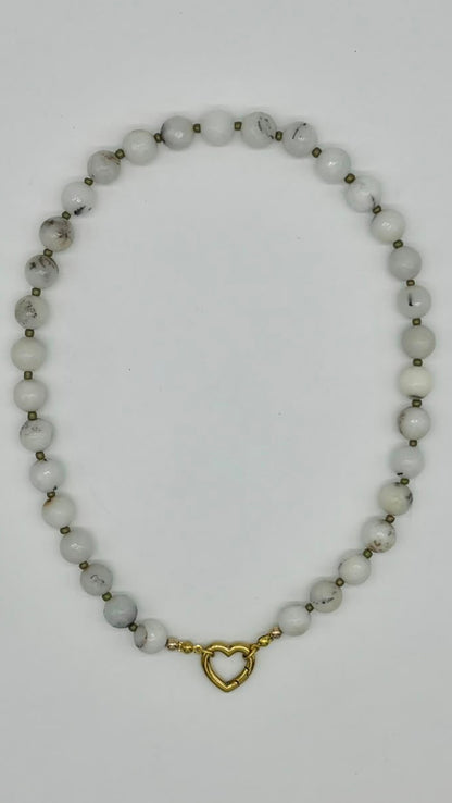 A beaded necklace made of small white beads with subtle gray marbling, separated by tiny metallic spacer beads, and finished with a small gold heart-shaped clasp, shown on a plain white background.