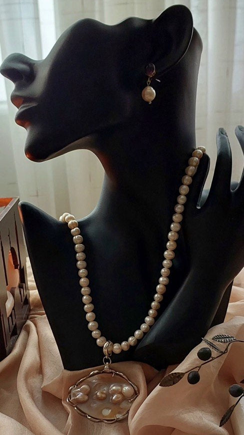 A pearl necklace with a large oval pendant and a matching pearl drop earring displayed on a black mannequin bust draped in pale pink fabric.