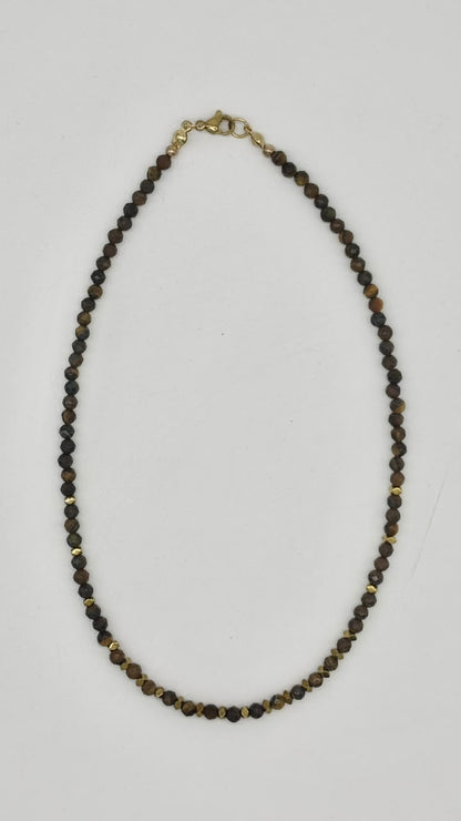 A beaded necklace made of small dark brown and black faceted beads with small gold spacer beads and a gold clasp, displayed on a light gray background.