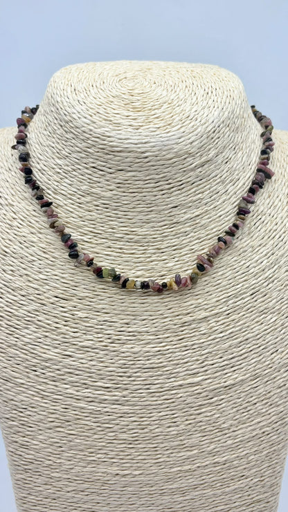 A beaded necklace made of small polished gemstone chips in earthy pink, green, brown, and black tones, displayed on a cream woven mannequin bust against a light blue background.