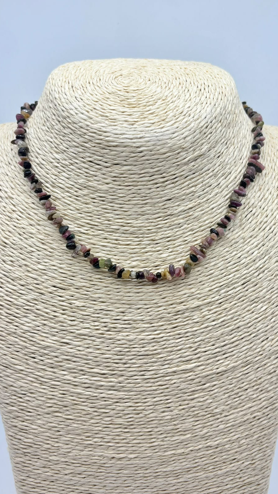 A beaded necklace made of small polished gemstone chips in earthy pink, green, brown, and black tones, displayed on a cream woven mannequin bust against a light blue background.