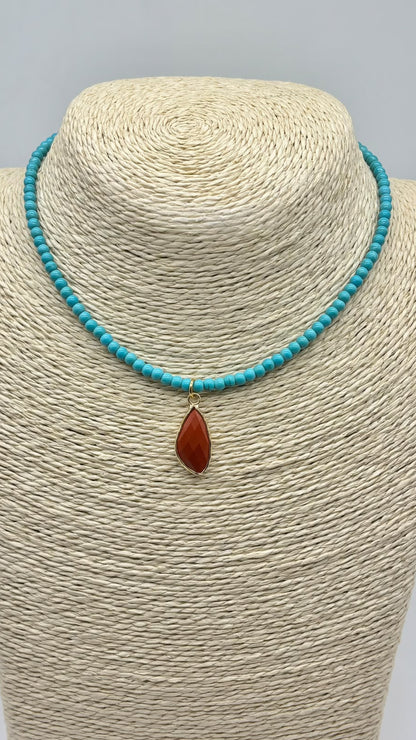 A turquoise beaded necklace with a teardrop-shaped reddish-brown stone pendant, displayed on a beige woven jewelry bust against a light gray background.