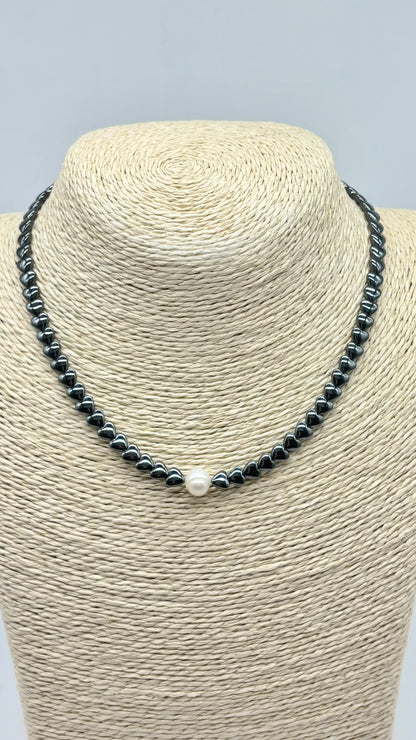 A beaded necklace displayed on a woven beige jewelry bust. The necklace consists of small dark gray beads with a single white pearl at the center front.