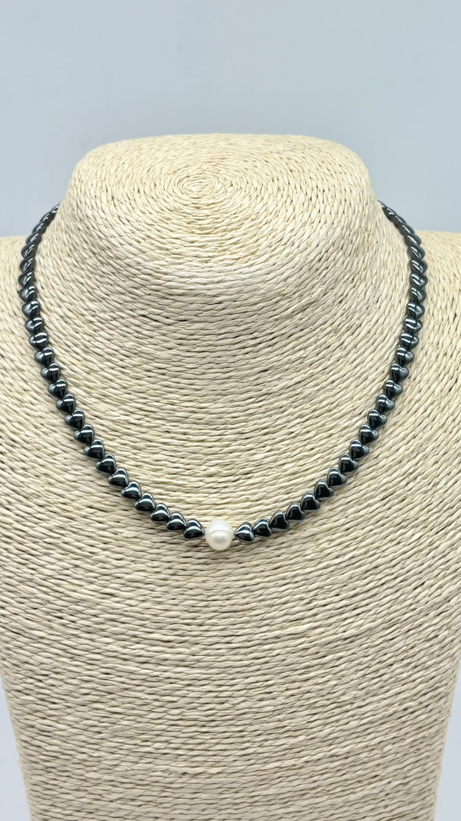 A beaded necklace displayed on a woven beige jewelry bust. The necklace consists of small dark gray beads with a single white pearl at the center front.