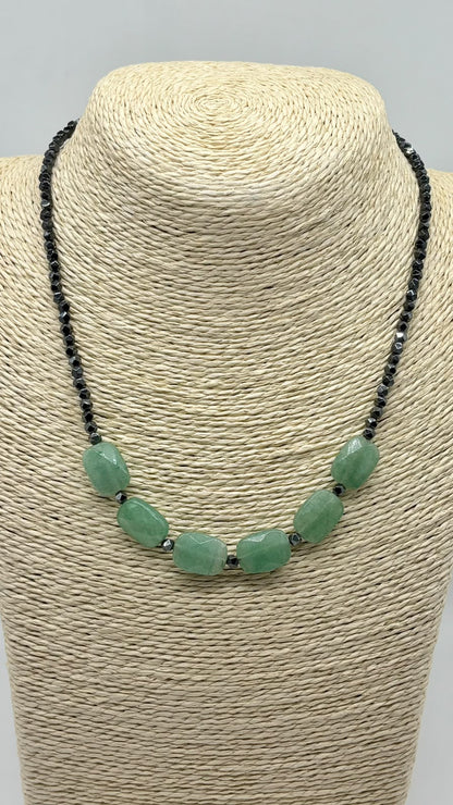 A statement necklace featuring seven polished light green jade-like oval beads strung on a black cord, displayed on a beige woven fabric jewelry bust against a light gray background.