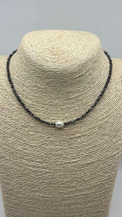 A beaded necklace with small dark gray beads and a single white pearl-like center bead, displayed on a woven beige jewelry bust against a light gray background.