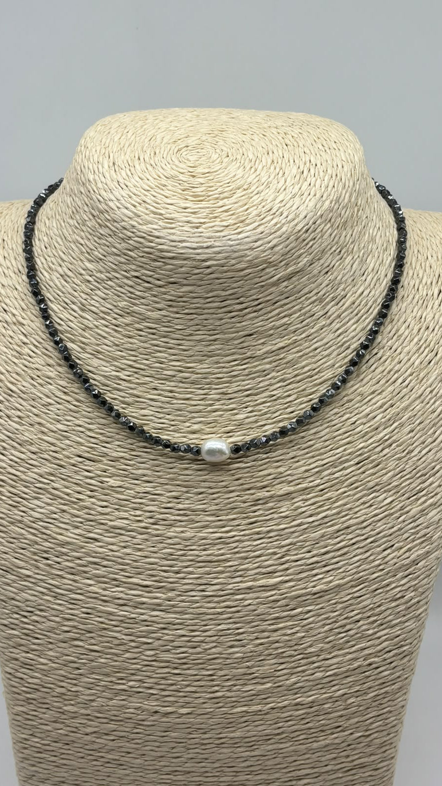 A beaded necklace with small dark gray beads and a single white pearl-like center bead, displayed on a woven beige jewelry bust against a light gray background.