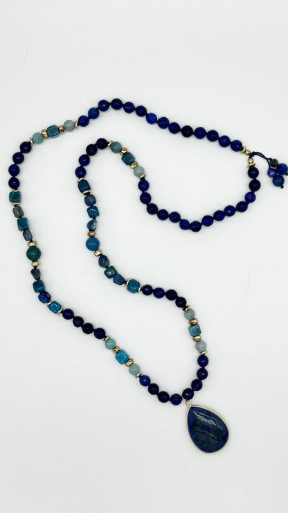 A beaded necklace featuring dark blue, navy, teal, and light blue stones, accented with small gold spacer beads, with a large dark blue teardrop pendant on a plain white background.