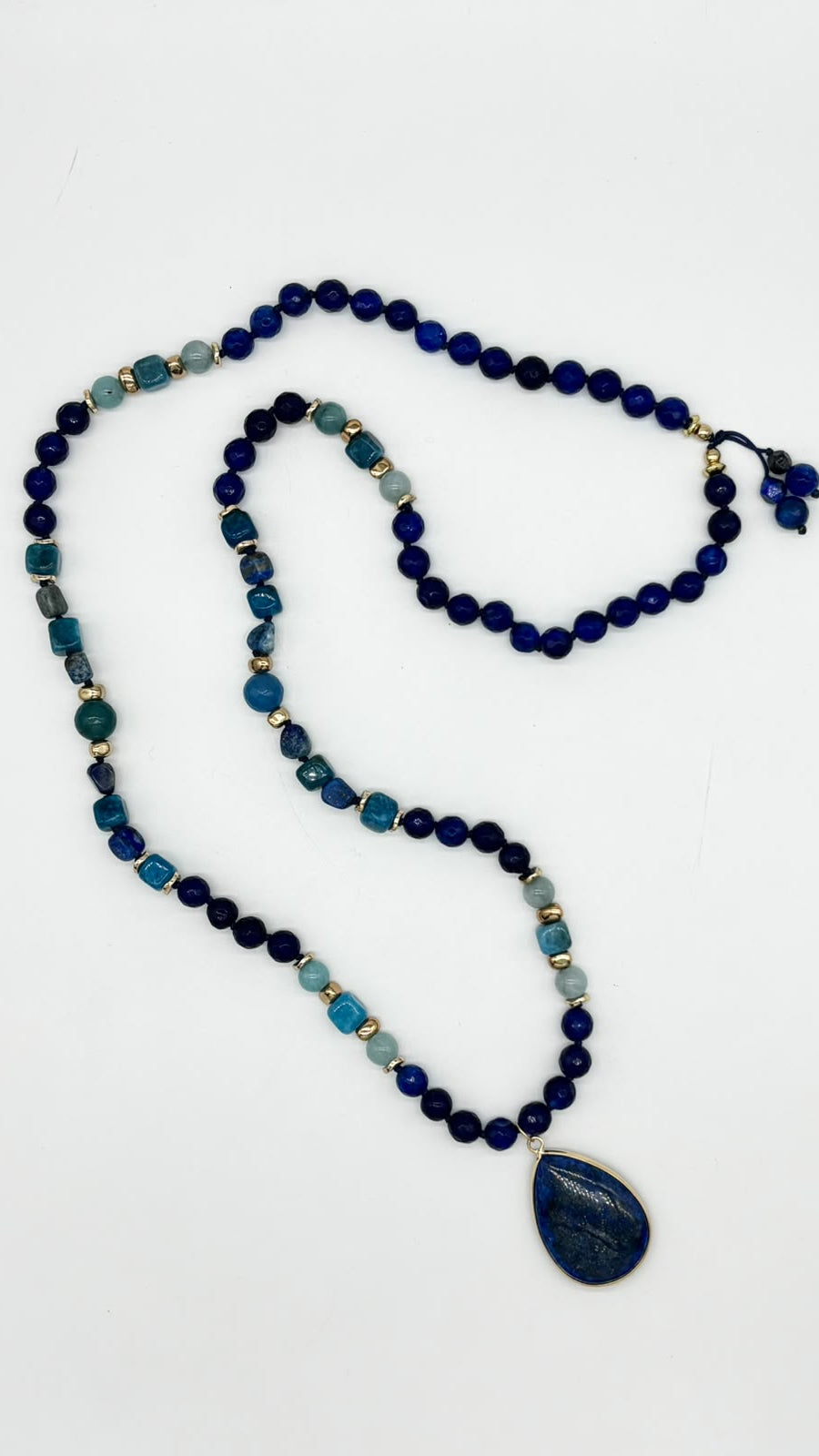 A beaded necklace featuring dark blue, navy, teal, and light blue stones, accented with small gold spacer beads, with a large dark blue teardrop pendant on a plain white background.