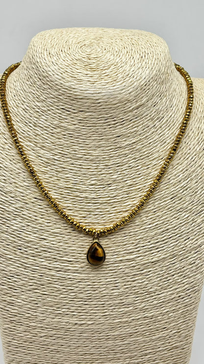 A gold-toned beaded necklace with a teardrop-shaped brown tiger's eye pendant, displayed on a beige woven mannequin bust against a light gray background.