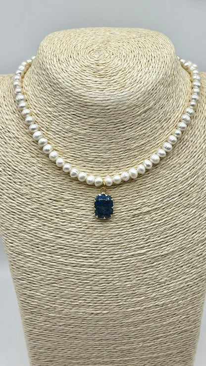 A pearl necklace with a small dark blue pendant, displayed on a beige woven jewelry bust against a light gray background.