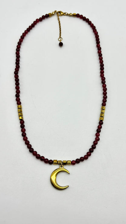 A beaded necklace featuring dark red round beads, gold spacer beads, and a gold crescent moon pendant, displayed on a plain white background.