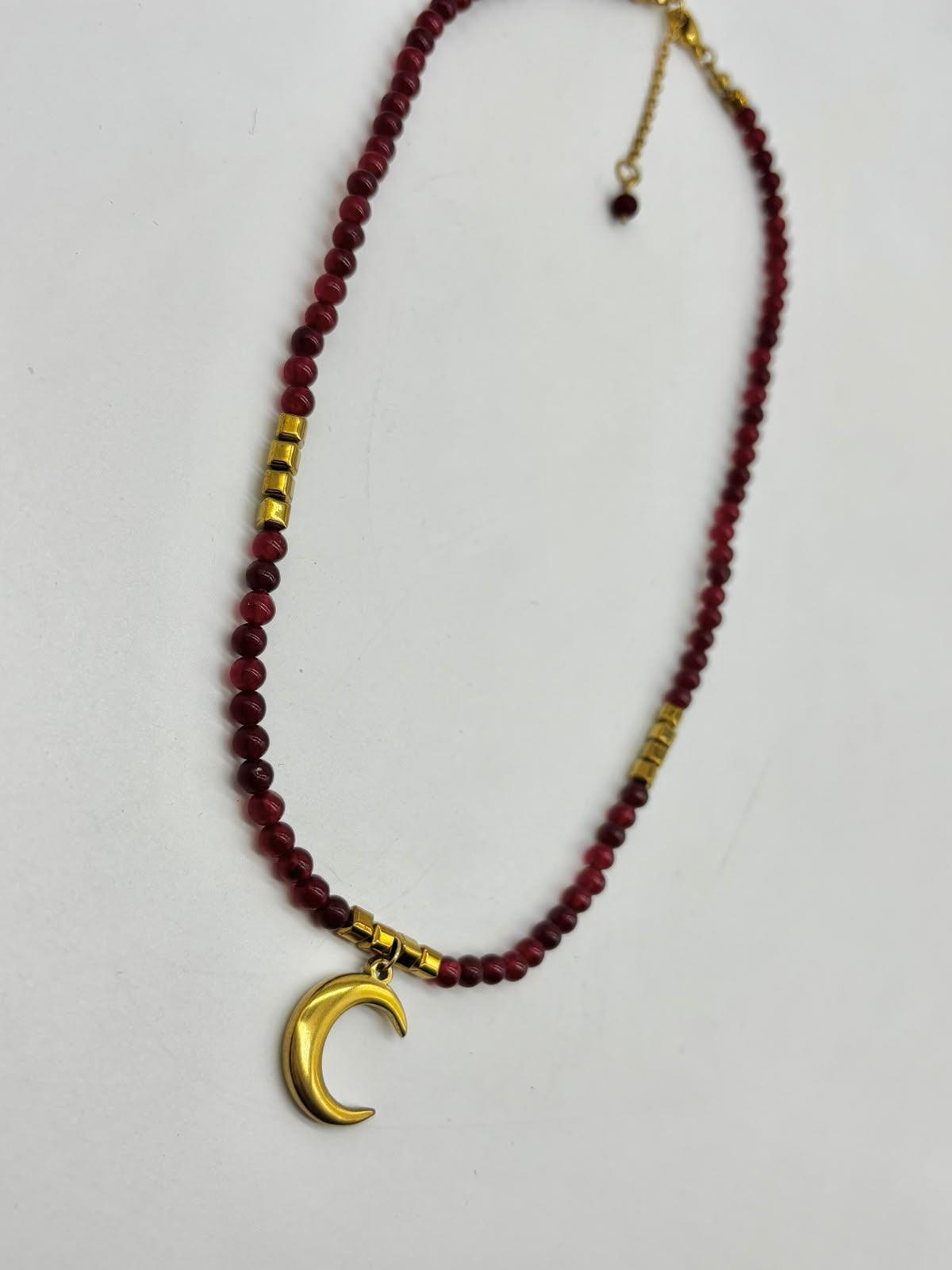 A beaded necklace featuring dark red faceted beads, small gold spacer beads, and a polished gold crescent moon pendant, displayed on a light gray background.