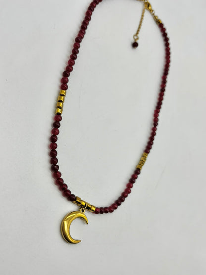 A beaded necklace featuring dark red faceted beads, small gold spacer beads, and a polished gold crescent moon pendant, displayed on a light gray background.