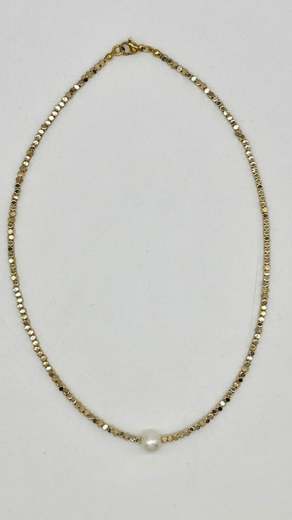 A delicate gold chain necklace with a single small white pearl bead at the center, laid flat on a light gray background.