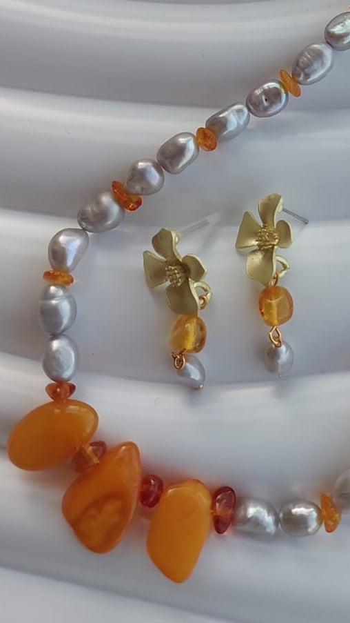 A beaded necklace and matching drop earrings displayed on a white wavy surface. The jewelry features white baroque pearls, small amber-orange beads, polished orange stone drops, and gold-tone metal hardware.