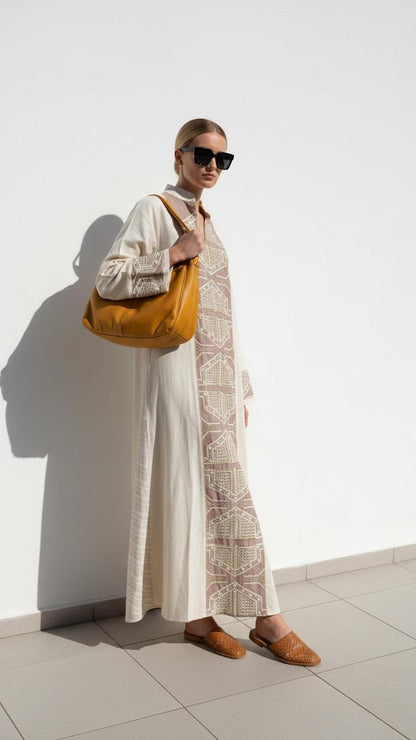 A woman stands against a white wall wearing a long cream dress with a beige geometric pattern panel, black sunglasses, and brown loafers, while carrying a large mustard yellow shoulder bag.