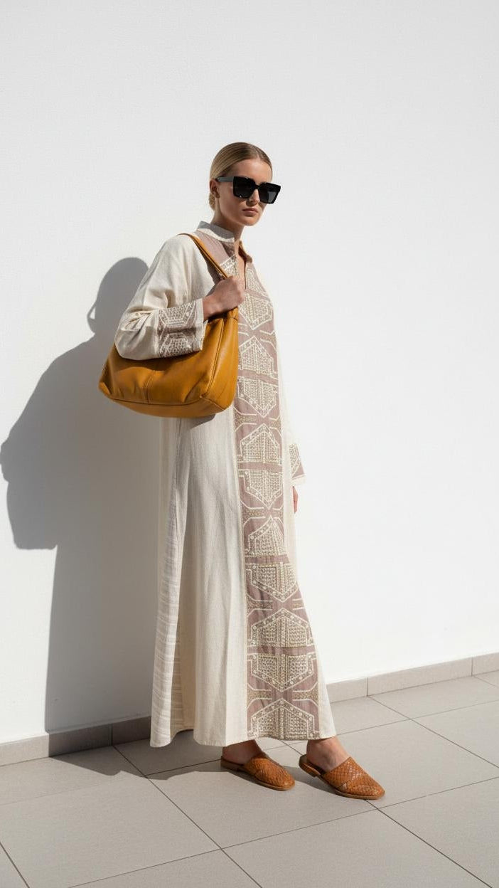 A woman stands against a white wall wearing a long cream dress with a beige geometric pattern panel, black sunglasses, and brown loafers, while carrying a large mustard yellow shoulder bag.