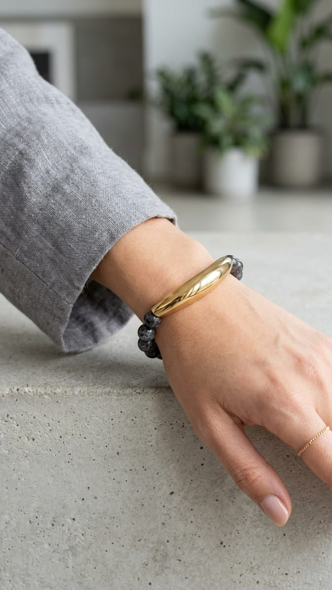 A person's hand resting on a concrete surface, wearing a polished gold bangle bracelet and a thin gold ring.