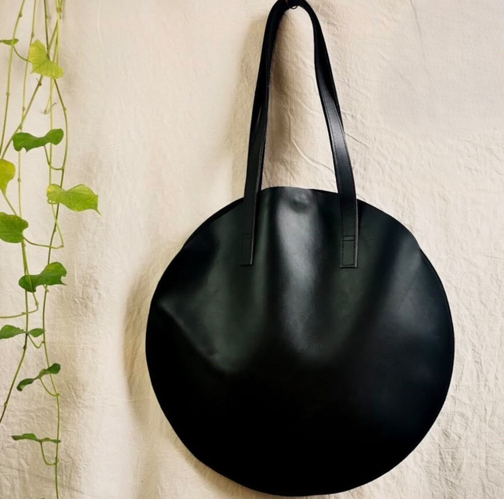 A large, round black leather tote bag with two shoulder straps, hanging from a metal hook against a textured off-white fabric backdrop with a green plant hanging on the left.