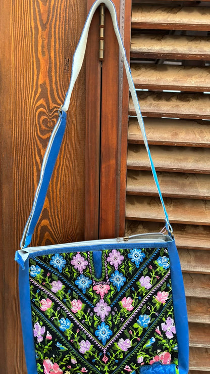 A blue and black embroidered shoulder bag with a floral pattern, hanging against a wooden slatted door.