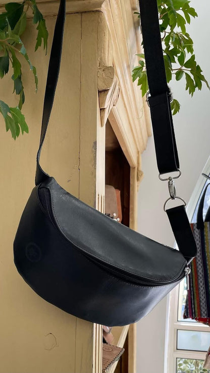 A black leather crescent-shaped shoulder bag with a wide strap, hanging against a beige wooden door with green leaves in the foreground.