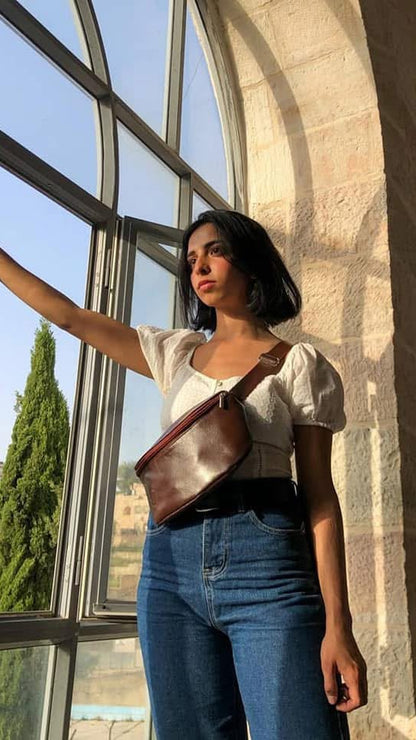 A woman stands by a large arched window in a sunlit stone building, wearing a white puff-sleeve top, blue jeans, and a brown leather crossbody bag.