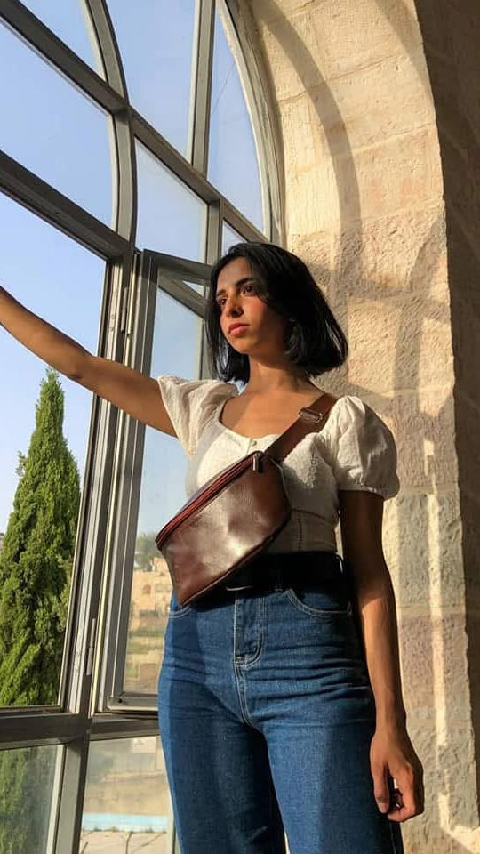 A woman stands by a large arched window in a sunlit stone building, wearing a white puff-sleeve top, blue jeans, and a brown leather crossbody bag.
