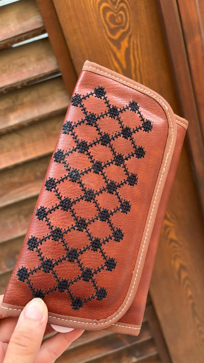 A hand holding a brown leather wallet with a black geometric diamond pattern, set against a background of wooden shutters.