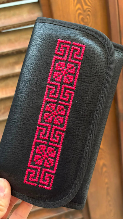 A black textured leather pouch with a bright pink geometric embroidered pattern, held by a hand in front of a wooden slatted door.