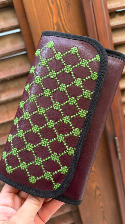 A hand holds a burgundy leather wallet or clutch with a green geometric patterned flap, set against a background of wooden shutters.