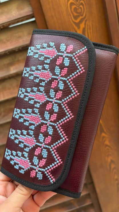 A hand holds a burgundy leather wallet with a colorful geometric embroidered pattern in pink and light blue on the front flap, set against a wooden background.