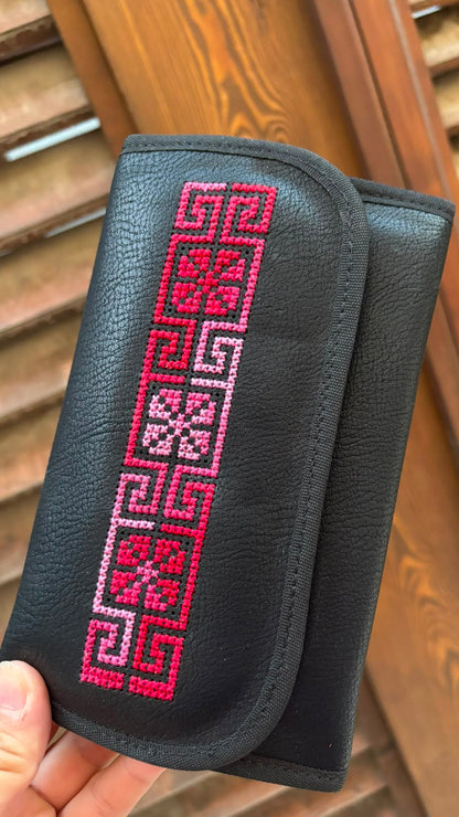 A black pebbled leather clutch wallet with a vertical pink and white embroidered geometric pattern, held by a hand in front of a wooden slatted background.