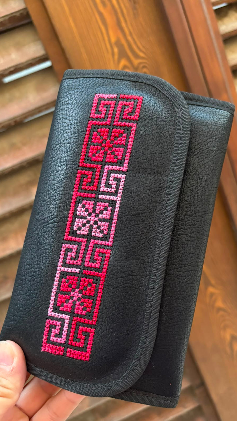 A black pebbled leather clutch wallet with a vertical pink and white embroidered geometric pattern, held by a hand in front of a wooden slatted background.
