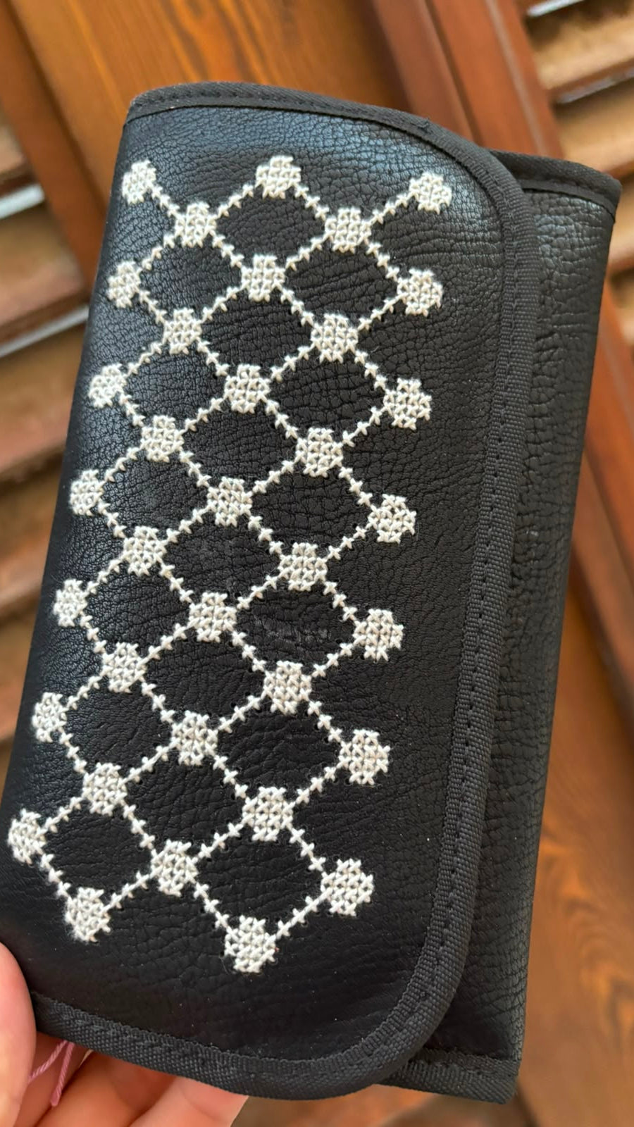 A black pebbled leather wallet or clutch with a white embroidered geometric diamond pattern, held by a hand in front of a wooden slatted background.