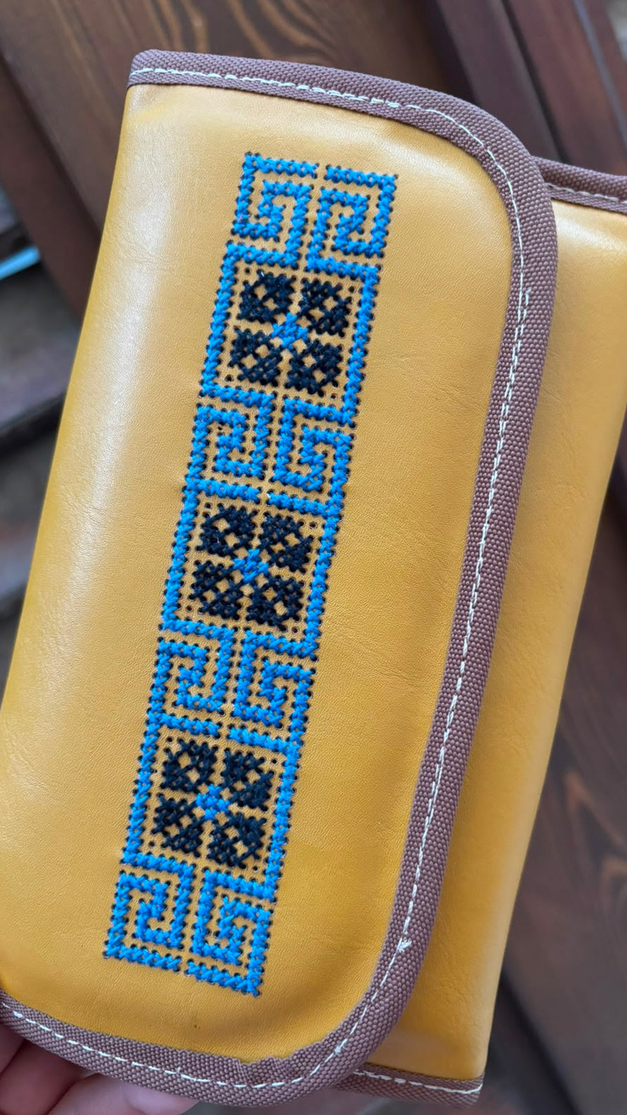 A mustard yellow leather wallet or clutch with a brown trim and a vertical embroidered geometric pattern in blue, black, and tan, held by a hand against a wooden background.