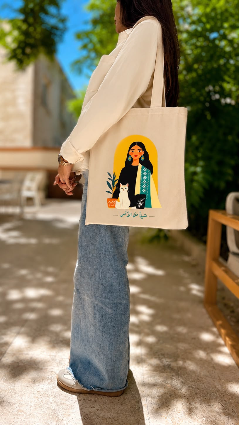 A woman in a cream long-sleeve top and light blue jeans carries a cream canvas tote bag with a colorful graphic illustration of a woman and a cat on a sunny outdoor patio.