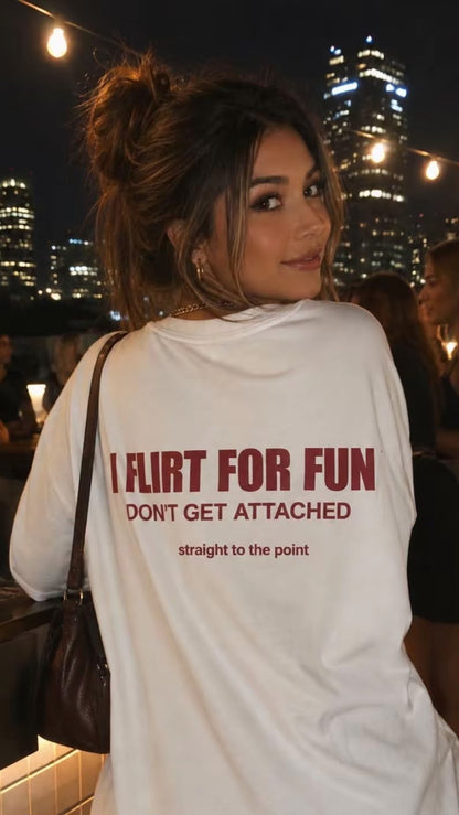 A woman at a night rooftop bar wearing a white oversized t-shirt with the text 'I FLIRT FOR FUN DON'T GET ATTACHED' printed in red on the back, with a city skyline and string lights in the background.