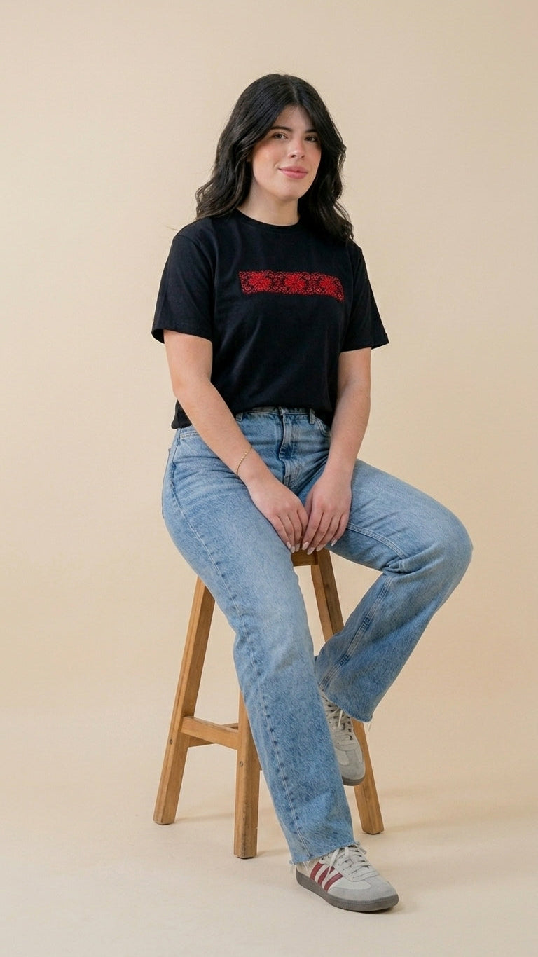 Woman sitting on a stool wearing a black t-shirt with red design and blue jeans against a beige background