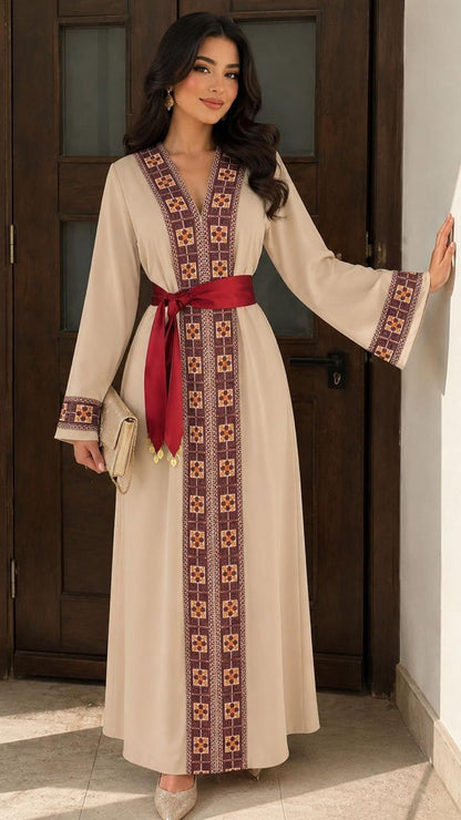A woman models a long beige kaftan dress with a red waist sash and intricate geometric embroidery in brown, red, and gold along the neckline, front panel, and cuffs. She is standing in a doorway with a wooden door and white walls in the background.
