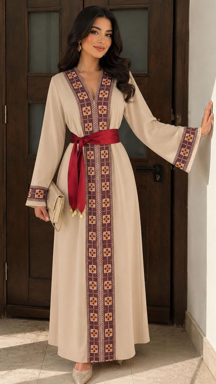 A woman models a long beige kaftan dress with a red waist sash and intricate geometric embroidery in brown, red, and gold along the neckline, front panel, and cuffs. She is standing in a doorway with a wooden door and white walls in the background.