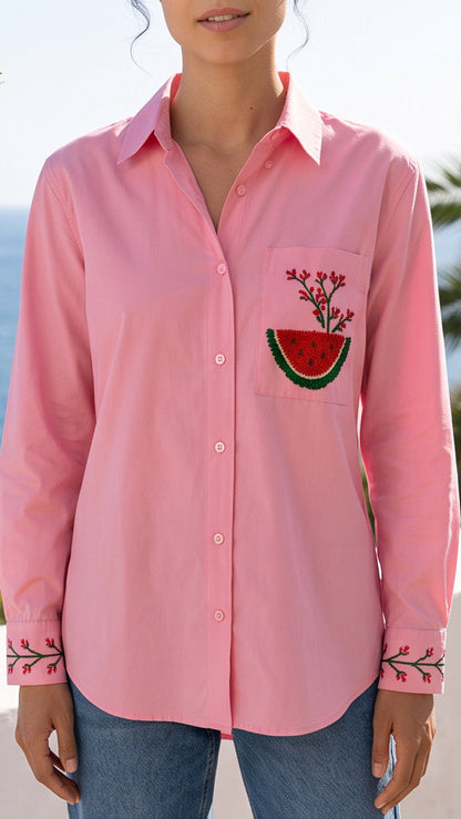 A woman wearing a pink long-sleeve button-up shirt with a watermelon patch embroidery on the pocket and floral embroidery on the cuffs, set against a sunny beach background with palm trees and the ocean.