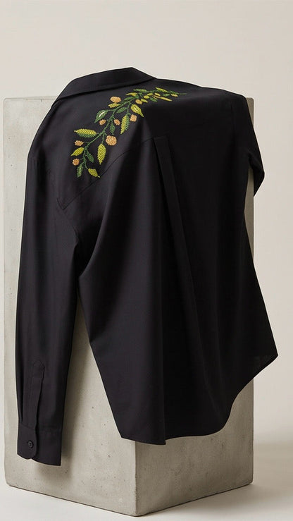 A black long-sleeve button-up shirt with a green and yellow floral leaf embroidery on the upper back, draped over a light gray concrete pedestal. The brand logo 'RONAQ' is visible in the top left corner.