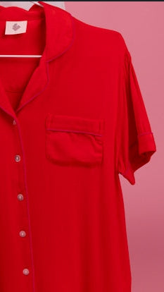 A bright red short-sleeve button-front shirt with a chest pocket, hanging on a white hanger against a solid pink background.