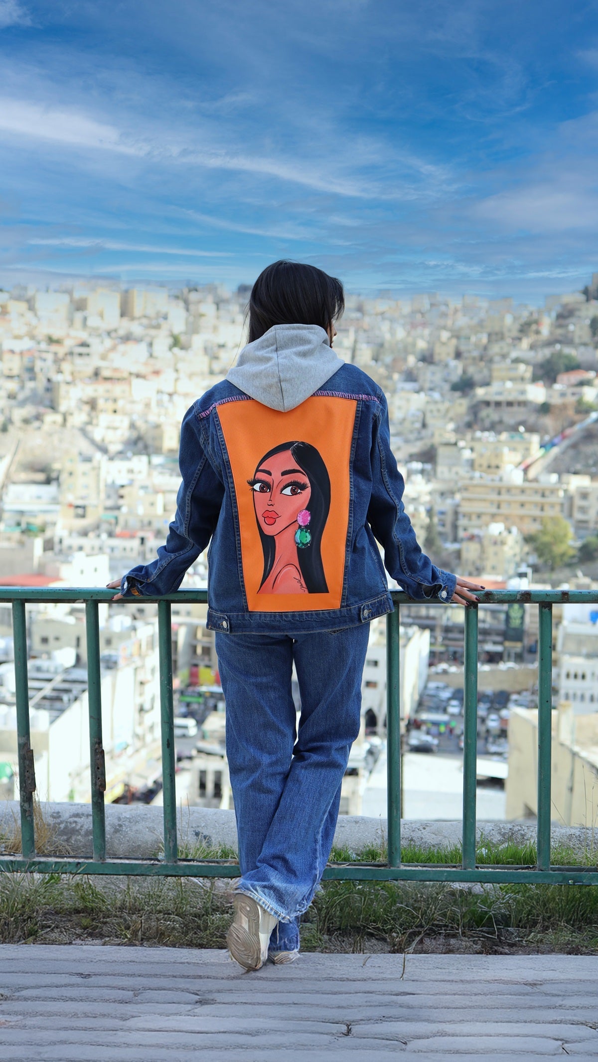 Person wearing a denim jacket with an orange back panel featuring a stylized face design, standing on a balcony overlooking a cityscape.