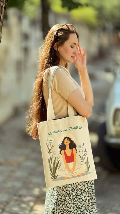 A woman carrying a cream canvas tote bag with Arabic text and an illustration of a woman meditating. The background is a softly blurred outdoor street scene.