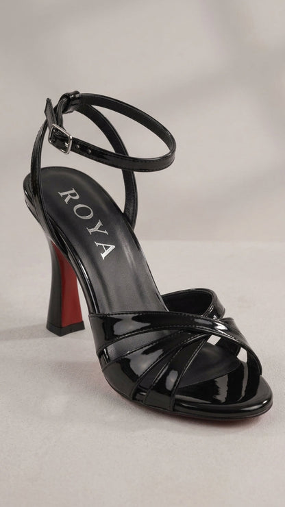 A black patent leather strappy sandal with an ankle strap, a high block heel with a red sole accent, and the brand name ROYA printed on the insole, set against a neutral light gray background.