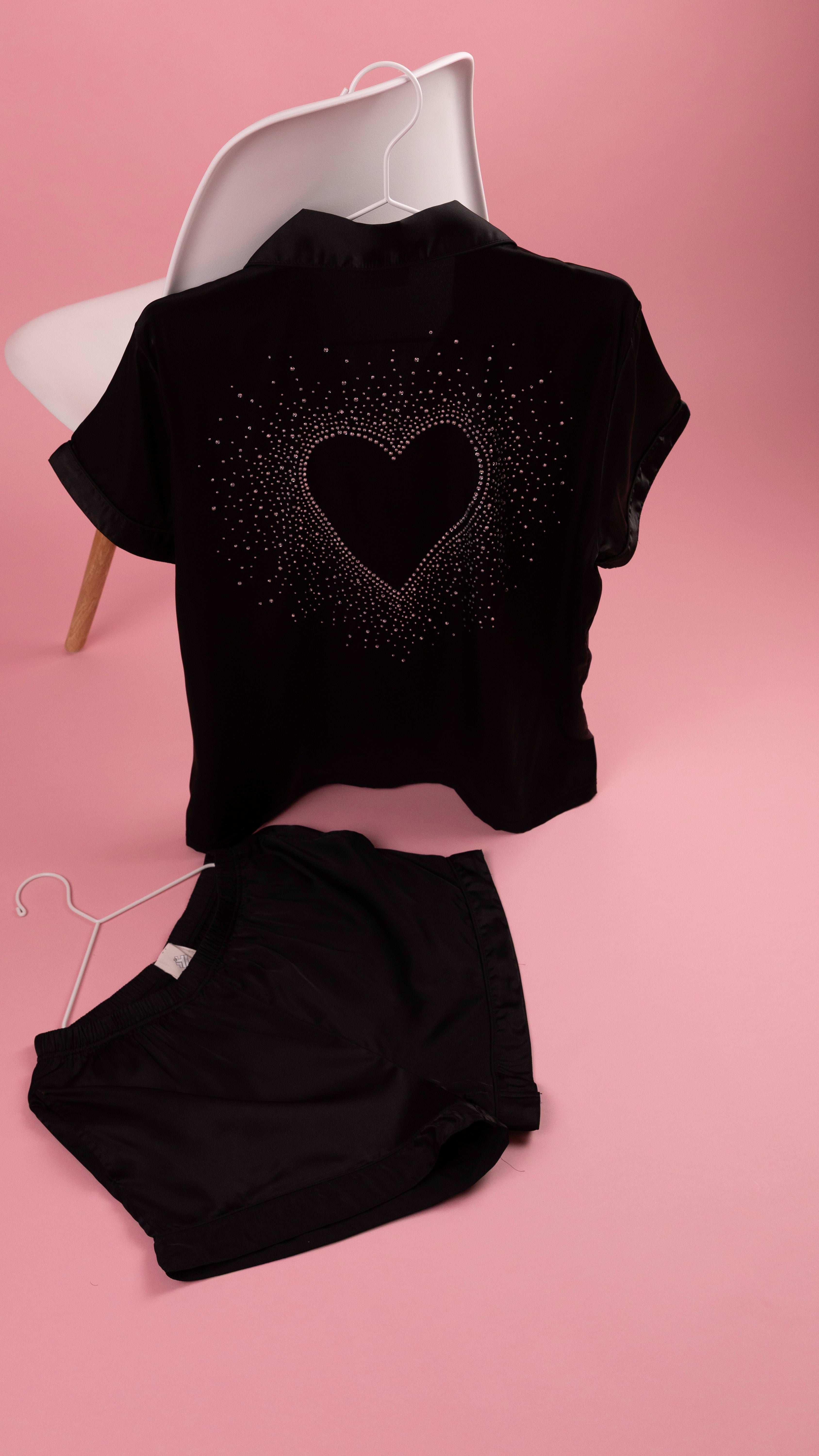 A black short-sleeve top and matching black shorts set displayed on hangers against a solid pink background. The top features a sparkling rhinestone heart design on the back.