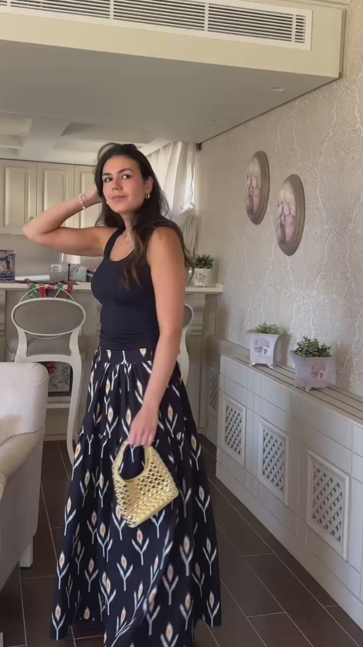Woman wearing a black sleeveless top and patterned long skirt holding a straw bag